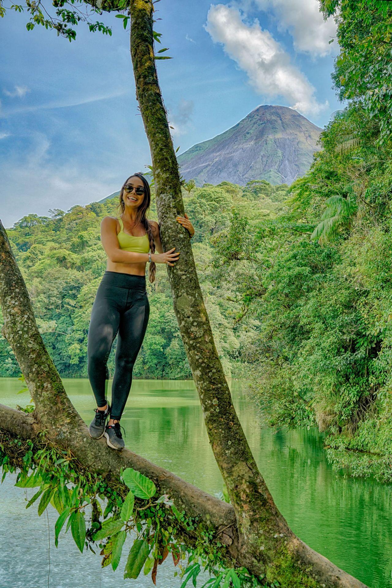 Woman standing on a tree branch by a lake with a mountain in the background.